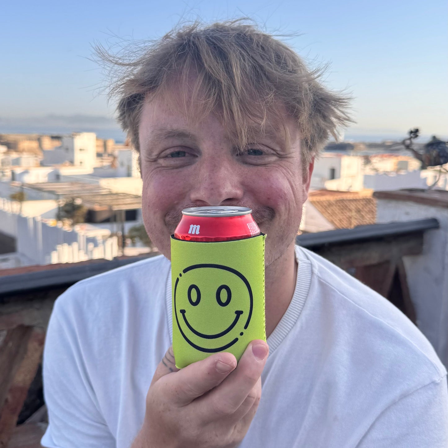 A man outdoors holding a red beverage can with a bright yellow smiley face can cooler. The background shows a clear blue sky and white buildings in the distance.