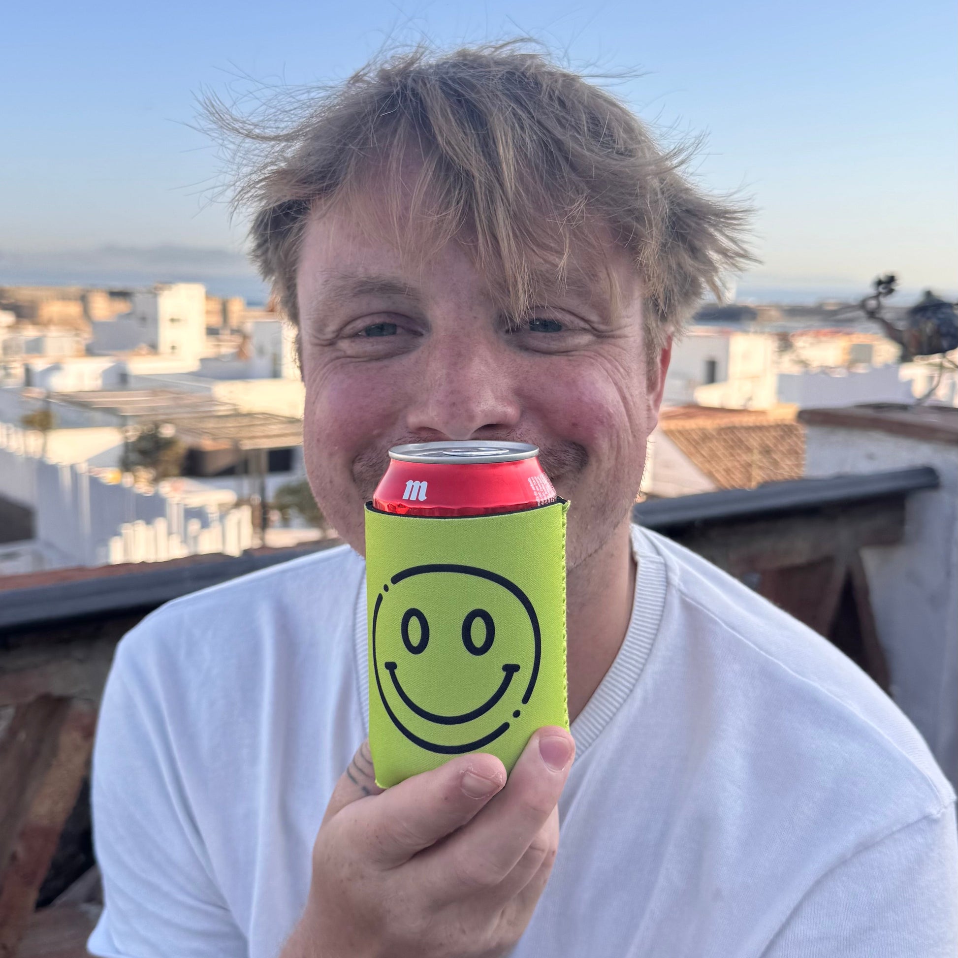 A man outdoors holding a red beverage can with a bright yellow smiley face can cooler. The background shows a clear blue sky and white buildings in the distance.
